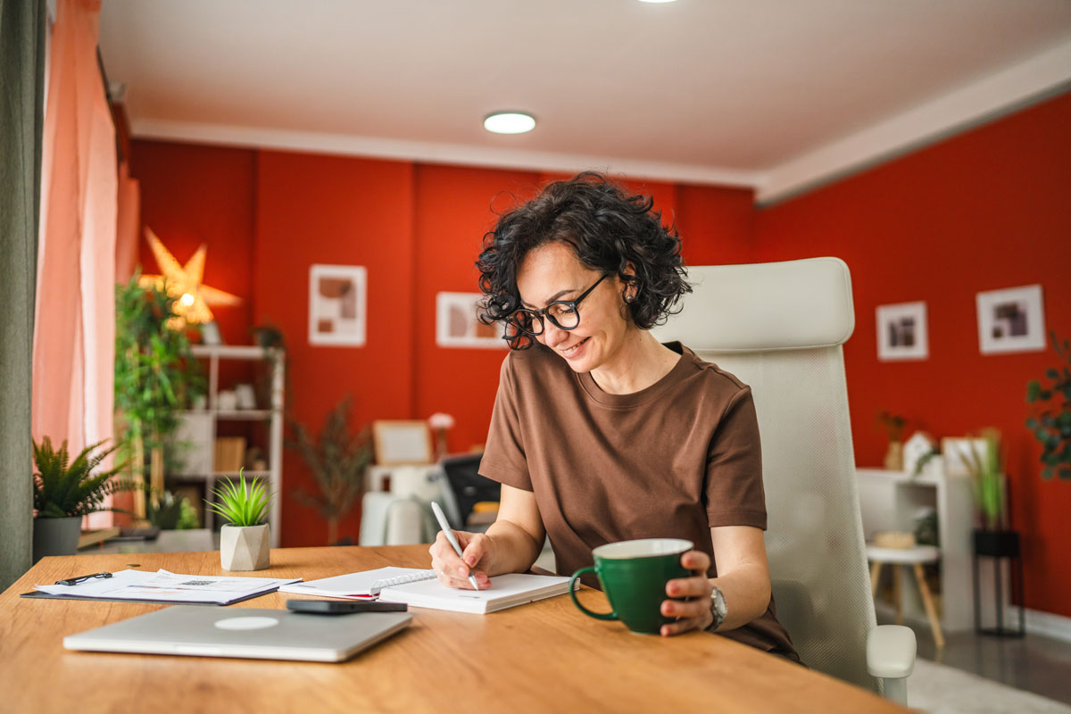 Woman sitting a desk writing in notebook with mug of coffee in her other hand